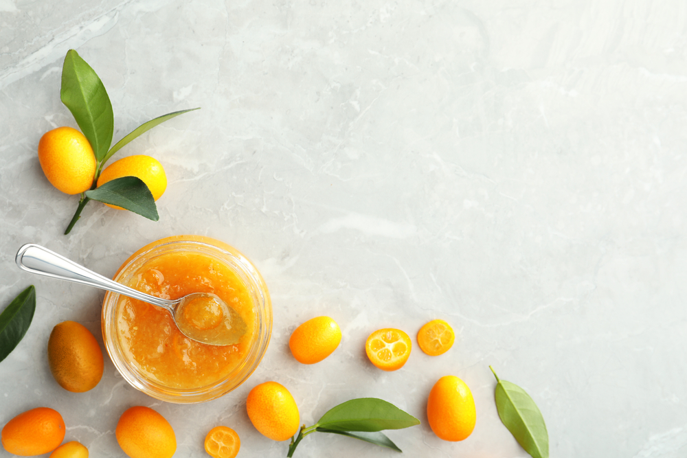 Fructe și frunze de kumquat pe o suprafață deschisă la culoare și un bol cu gem realizat din aceste fructe.