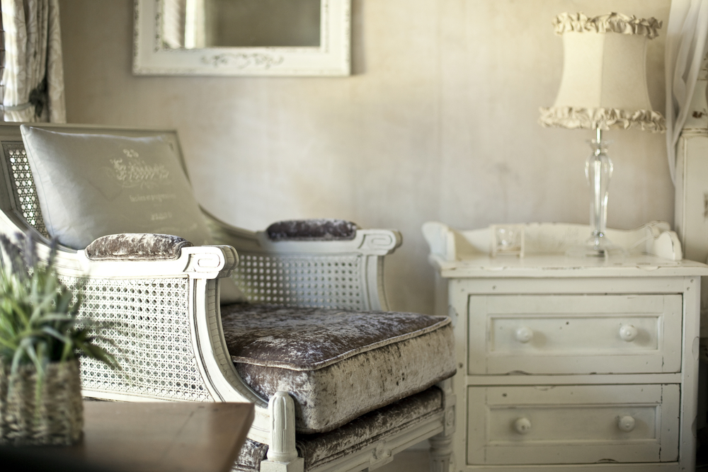 Un colț de dormitor în stilul shabby chic cu un fotoliu în nuanțe neutre și noptieră albă cu vopsea scorojită.