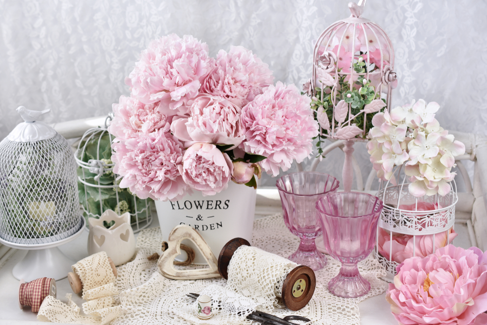 Decorațiuni shabby chic, cum ar fi dantelă albă, vază din metal cu bujori roz proaspeți, veselă pastelată și inimioară din lemn.