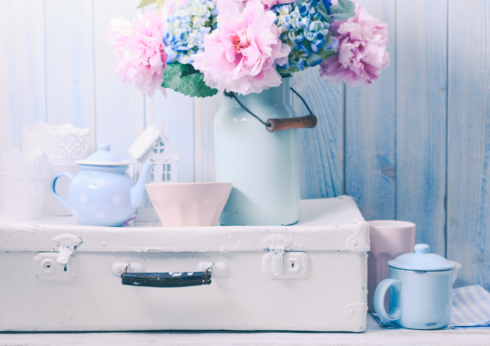 Decor în stilul shabby chic în naunțe pastelate cu o valiză albă vintage și buchet de flori roz și bleu.