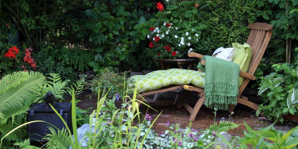 Un șezlong din lemn natur printre plante cu flori într-0 grădină mică.