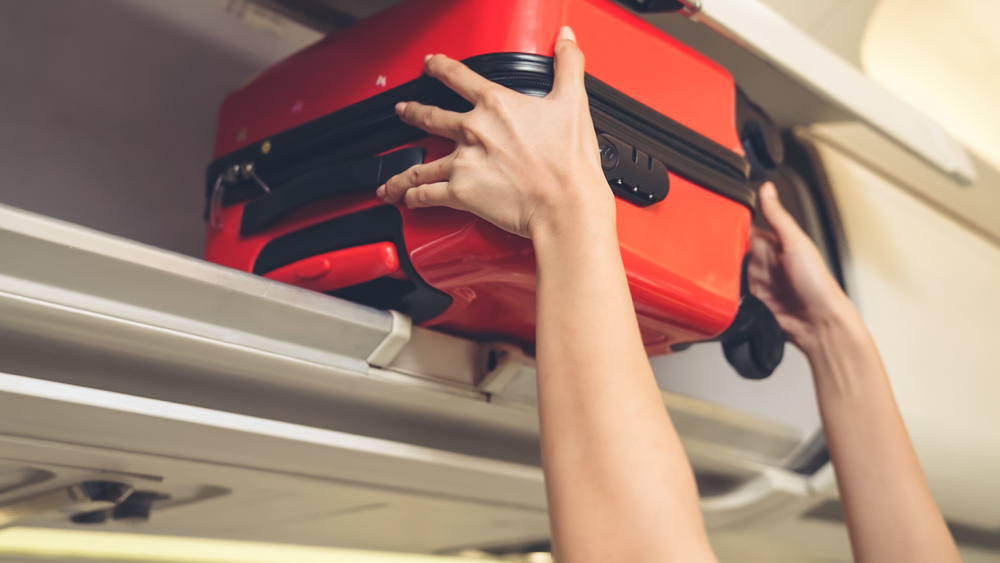 Mâini de femeie pun în spațiul pentru bagaje din avion un bagaj mic roșu, cu margini negre.