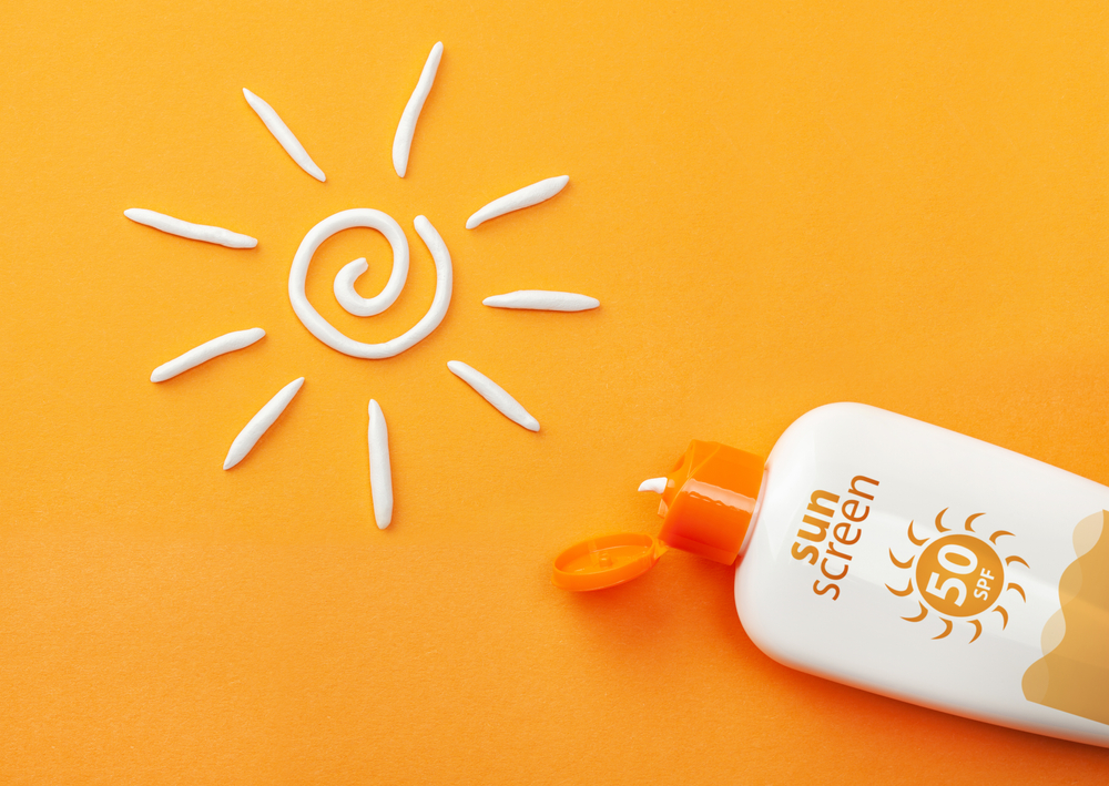 Loțiune SPF de pus în bagajul pentru mare: o loțiune cu factor de protectie 50 pe un fundal portocaliu și cu soare cu raze desenat cu cremă albă