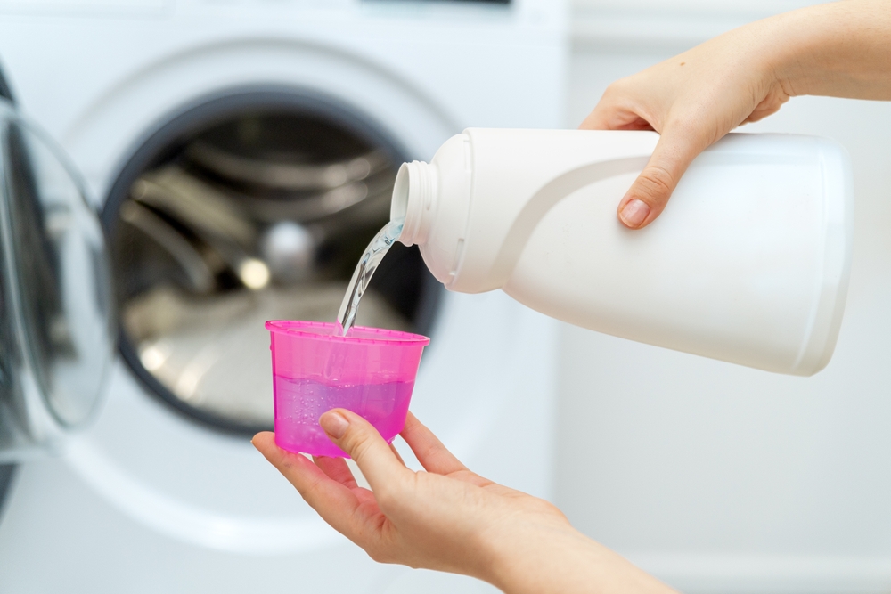 Mâini de femeie pun balsam de rufe sau detergent lichid într-un recipient roz, în fața mașinii de spălat haine.