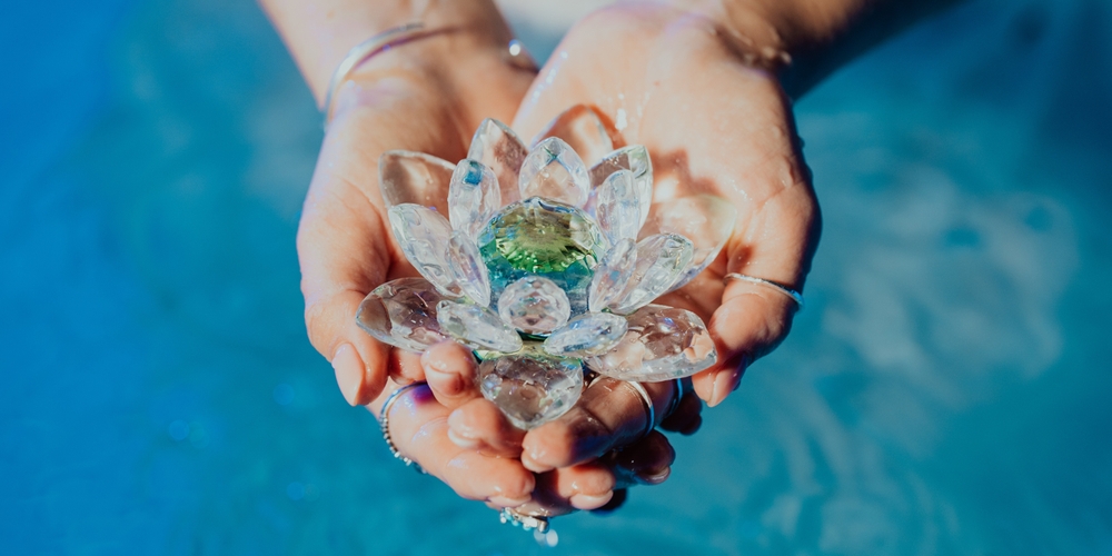 Mâini de femeie țin un lotus de cristal deasupra unei ape albastre.