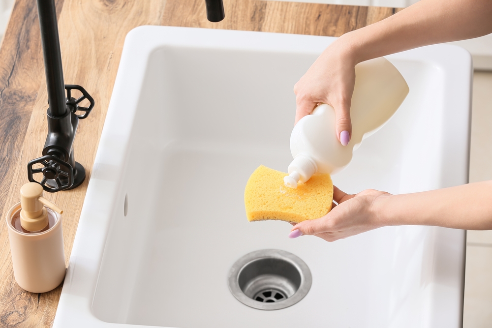 Soluție curățare baterii negre: Mâini de femeie pun pe un burete galben detergent de vase, deasupra unei chiuvete albe cu baterie neagră.