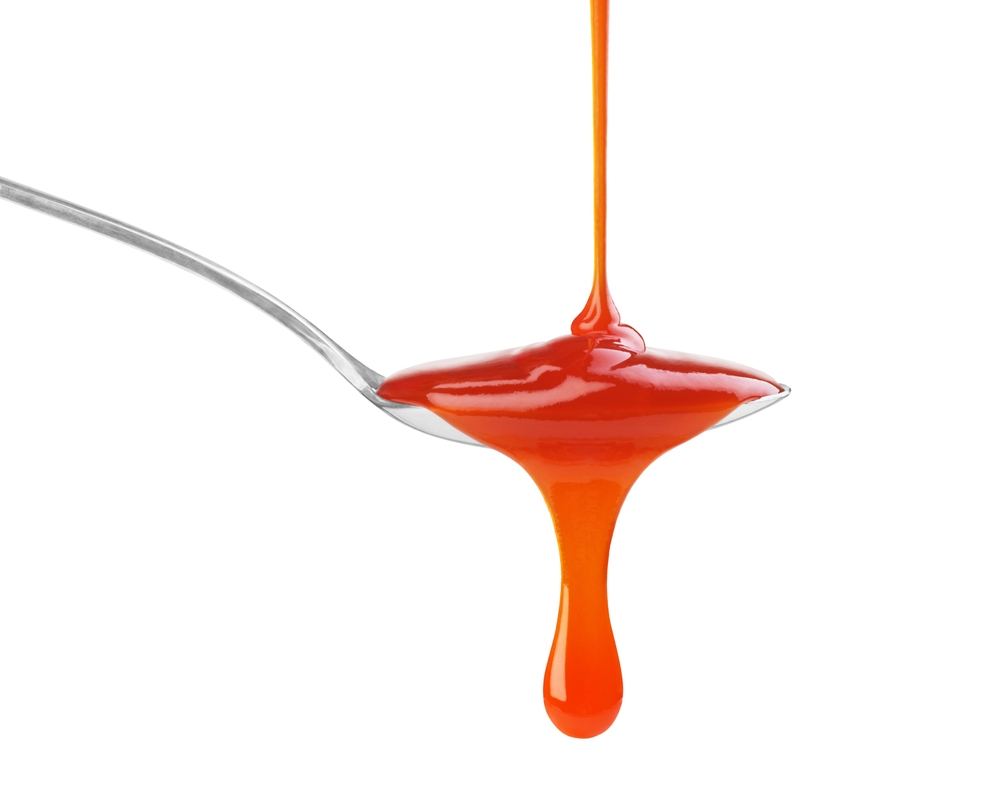 O lingură din argint în care curge ketchup, pe un fundal alb.