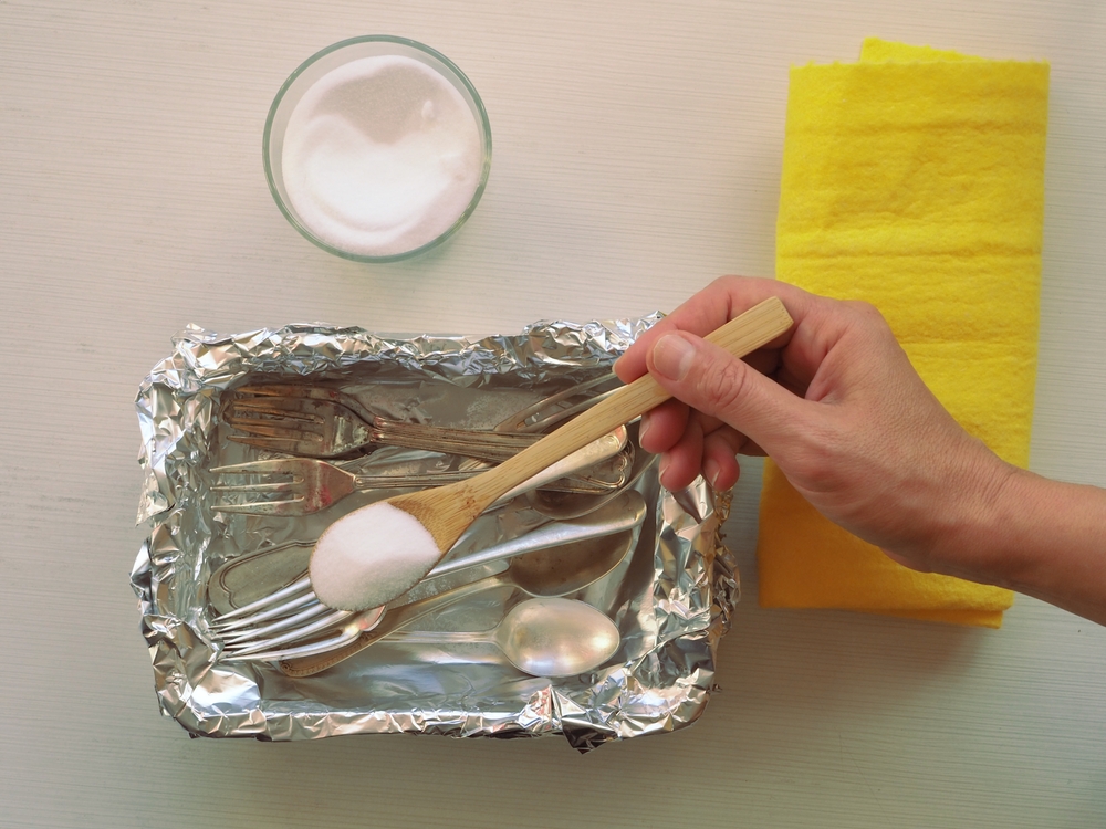 Curățare argint cu folie de aluminiu: O mână pune bicarbonat de sodiu sau sare într-un recipient din folie de aluminiu cu tacâmuri din argint, pe o suprafață albă, alături de o lavetă galbenă și un bol mic cu un praf alb.
