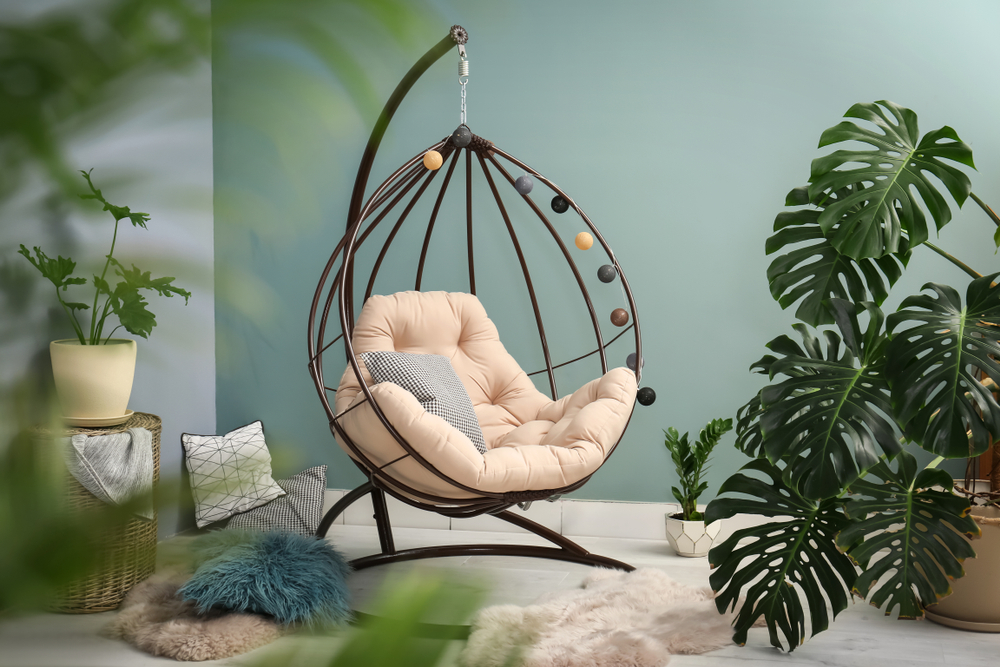 Plante interior mari: O plantă monstera într-o amenajre cu mochetă albă, pereți verde deschis și balansoar de o persoană cu perne decorative crem deschis.