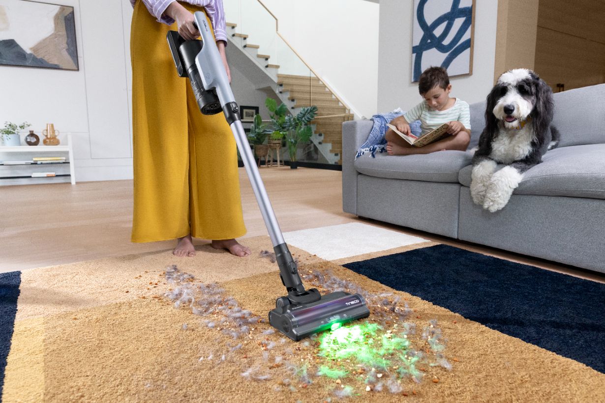 femeie foloseste un aspirator tineco pure one station 5 pentru a inlatura mizeria de pe covor in timp ce un copil si un catel stau pe canapea